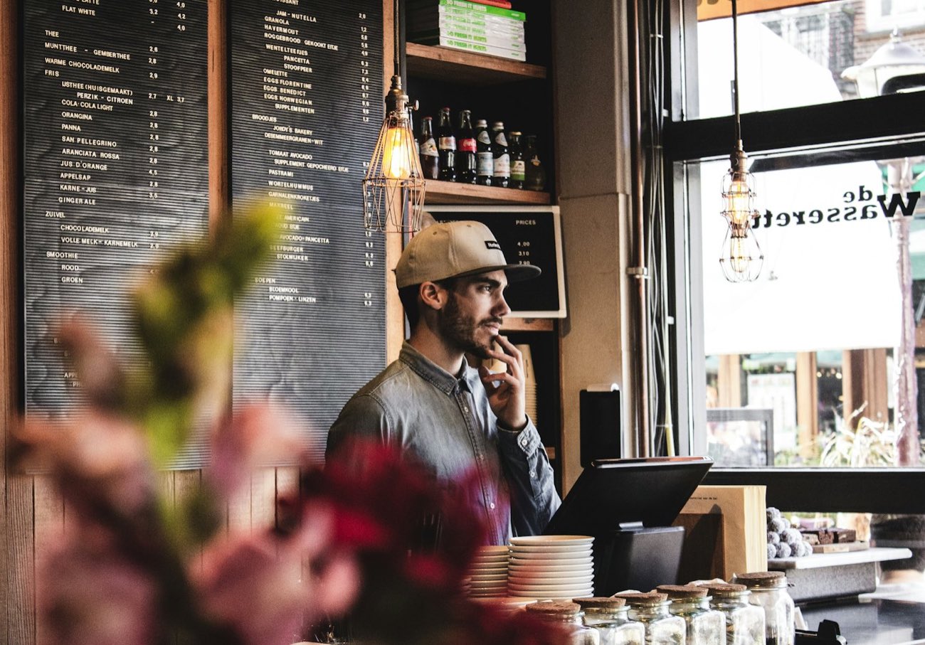 A café owner behind the counter, reading, hand on his chin.